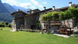 A stone building with a wooden deck and a picnic table, surrounded by a well-maintained garden and a mountain backdrop. - Le Case dei Baff (Ardenno)