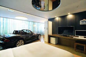 A modern hotel room with a convertible sports car, a flat-screen TV, and a compact refrigerator.