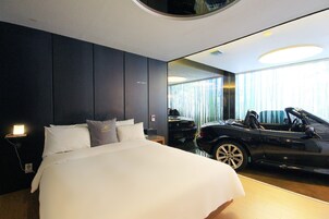 A modern hotel room with a large bed, a car in the background, and a view of the city through large windows.