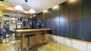 Una zona de bar con mostrador de madera y dos taburetes, un refrigerador y estanterías con botellas.