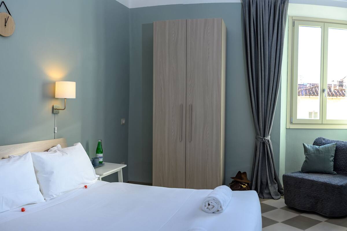 A hotel room with a bed, bedside tables, a mirror, a clock, a wardrobe, and a window with curtains.