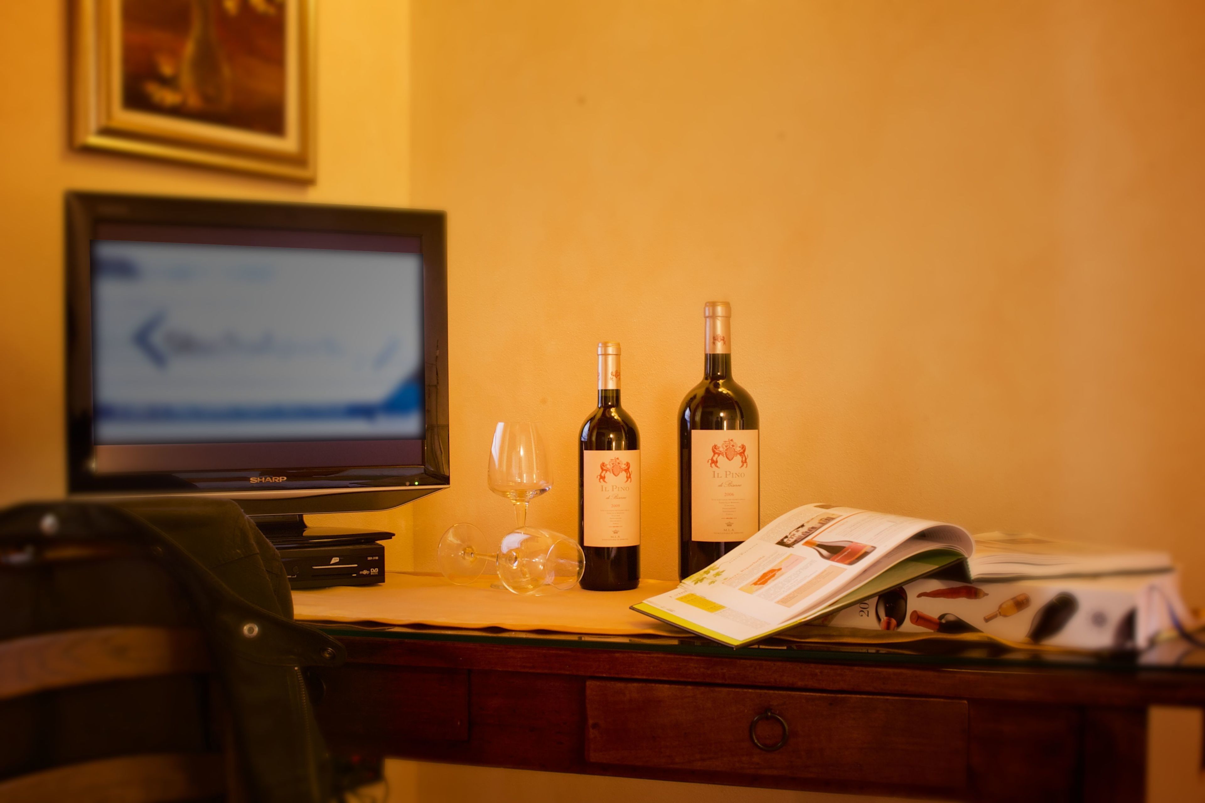 Una scrivania con un monitor del computer, due bottiglie di vino, un bicchiere da vino e una rivista.