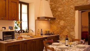 Une cuisine avec des meubles en bois, un mur en pierre, un coin repas avec une table dressée pour deux personnes et une fenêtre avec des rideaux.