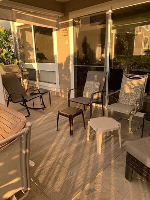 Een zonnig terras met een houten tafel, meerdere stoelen en een klein krukje.