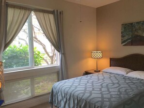 A hotel room with a bed, a nightstand, a lamp, a window with curtains, and a painting on the wall. - Oceanview Townhome w/ Pool Near Beaches & Shops (Kailua-Kona)