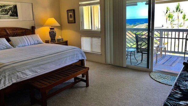 A hotel room with a bed, a nightstand, a lamp, a window with blinds, and a balcony with a view of the ocean.