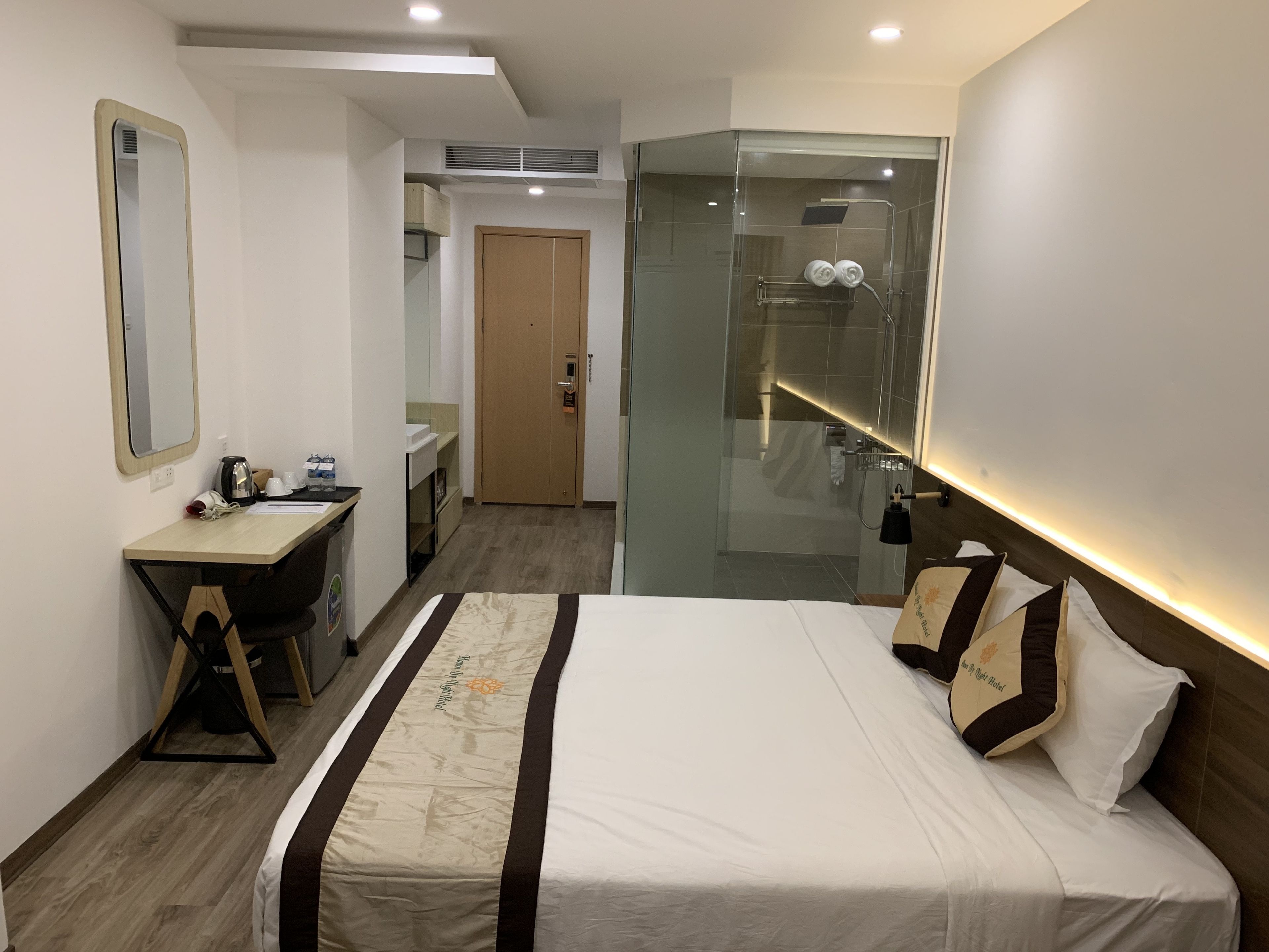 A hotel room with a bed, a desk, a mirror, a shower, and a door.