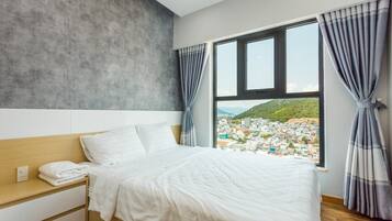A hotel room with a large bed, a wooden headboard, a nightstand, and a view of a cityscape through a window with curtains.