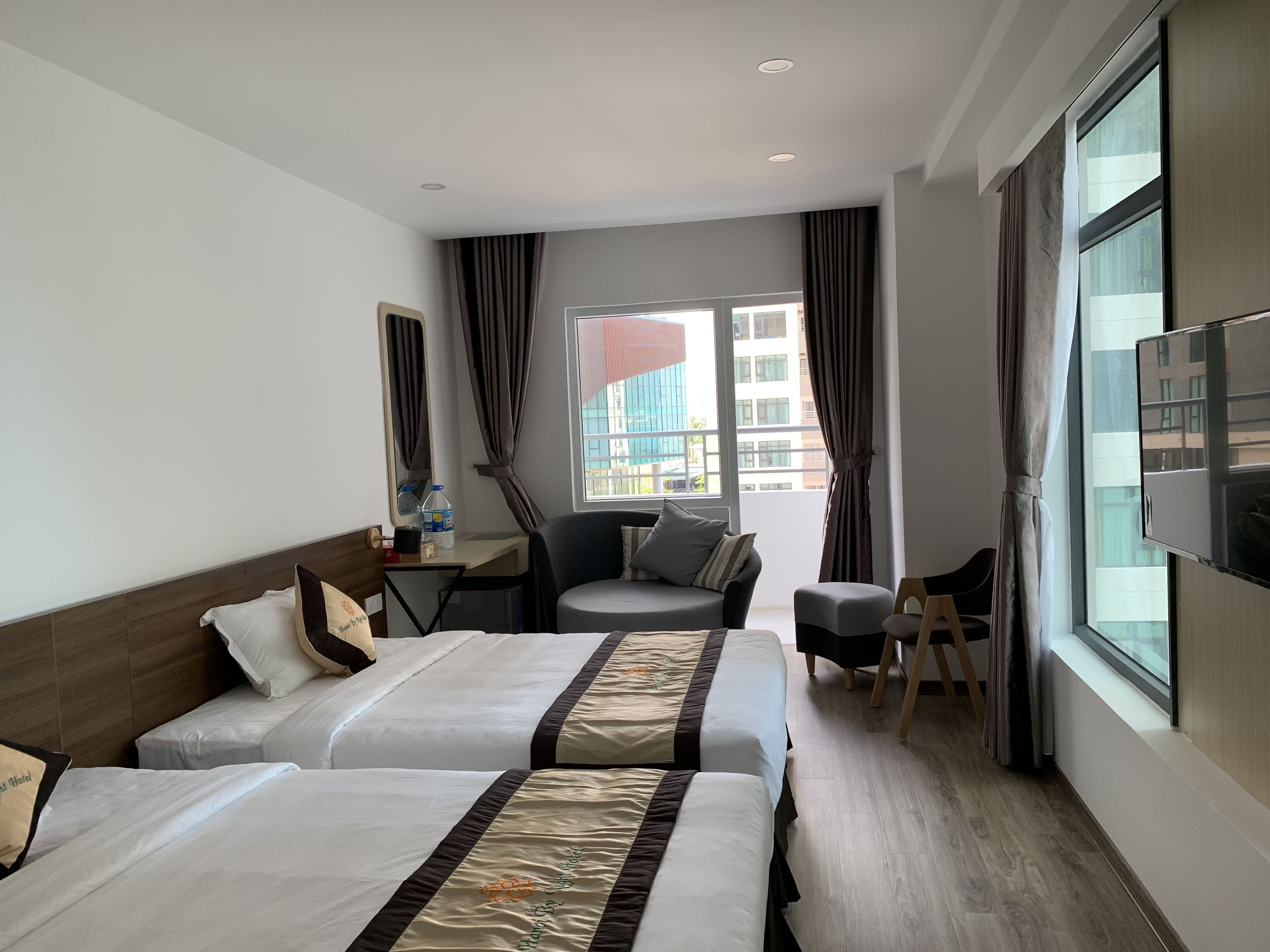 Quarto de hotel com duas camas, uma escrivaninha, uma cadeira, televisão e uma janela grande com cortinas.
