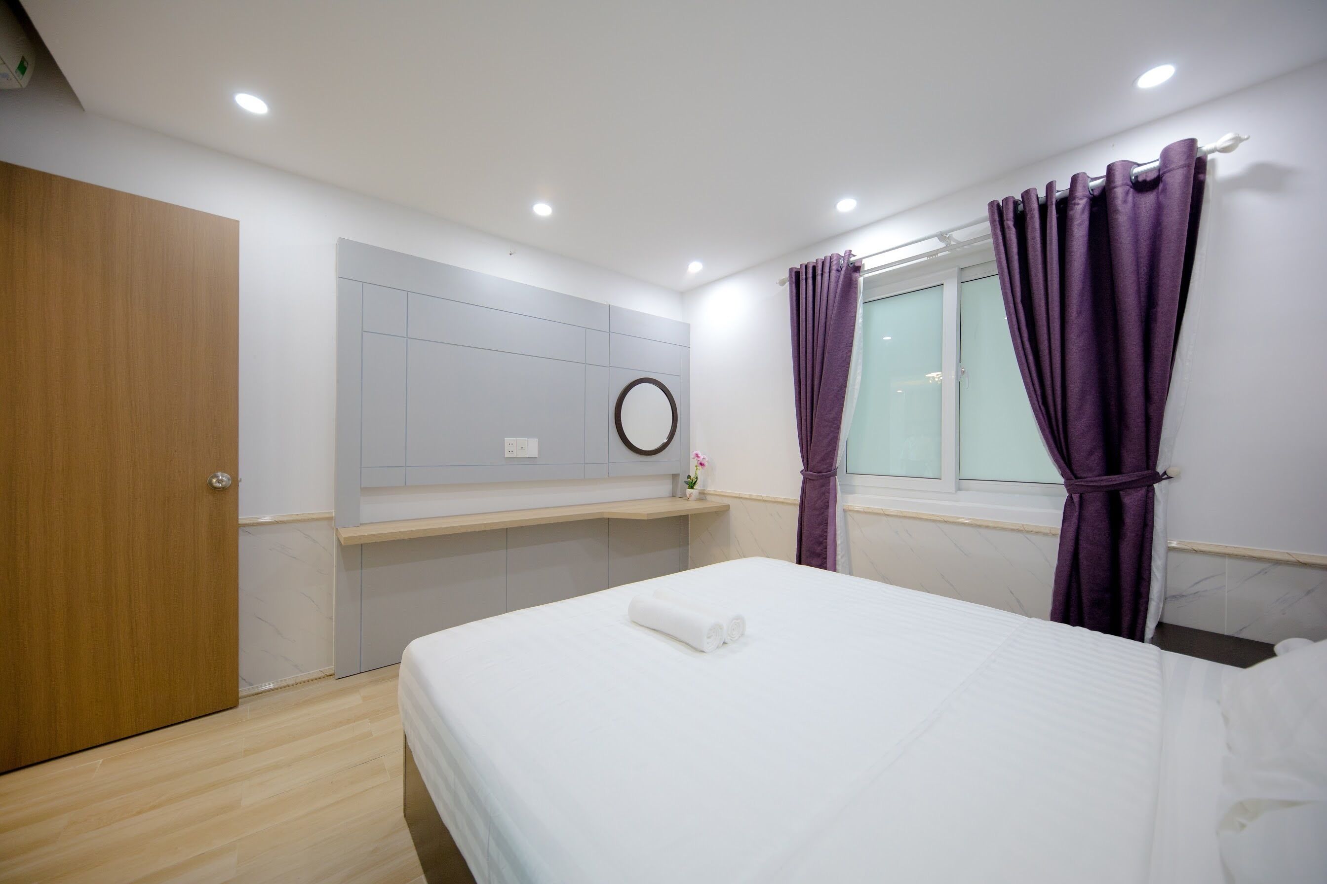 Habitación de hotel con cama, puerta de madera, paredes blancas, ventana con cortinas moradas y un espejo redondo pequeño en la pared.
