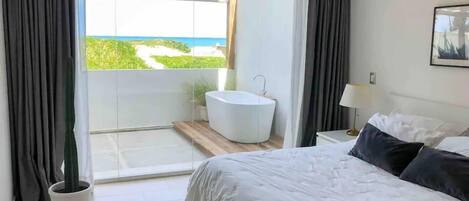 A modern bedroom with a large bed, a bathtub, and a balcony with a view of the sea.