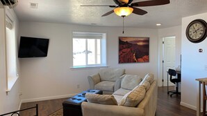 Una sala de estar con ventilador de techo, un televisor de pantalla plana, una ventana, un cuadro y un reloj en la pared.