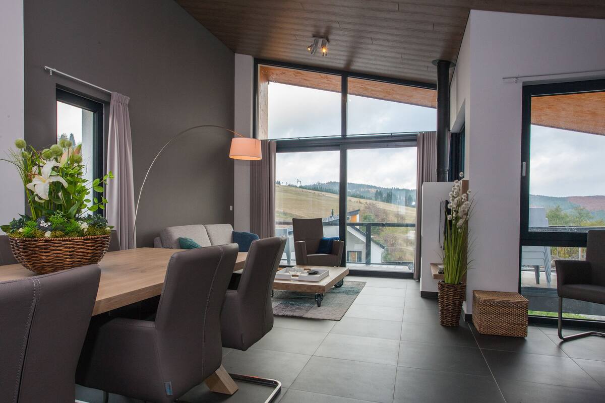 A modern dining area with a wooden table, grey upholstered chairs, and a large window offering a view of the countryside.