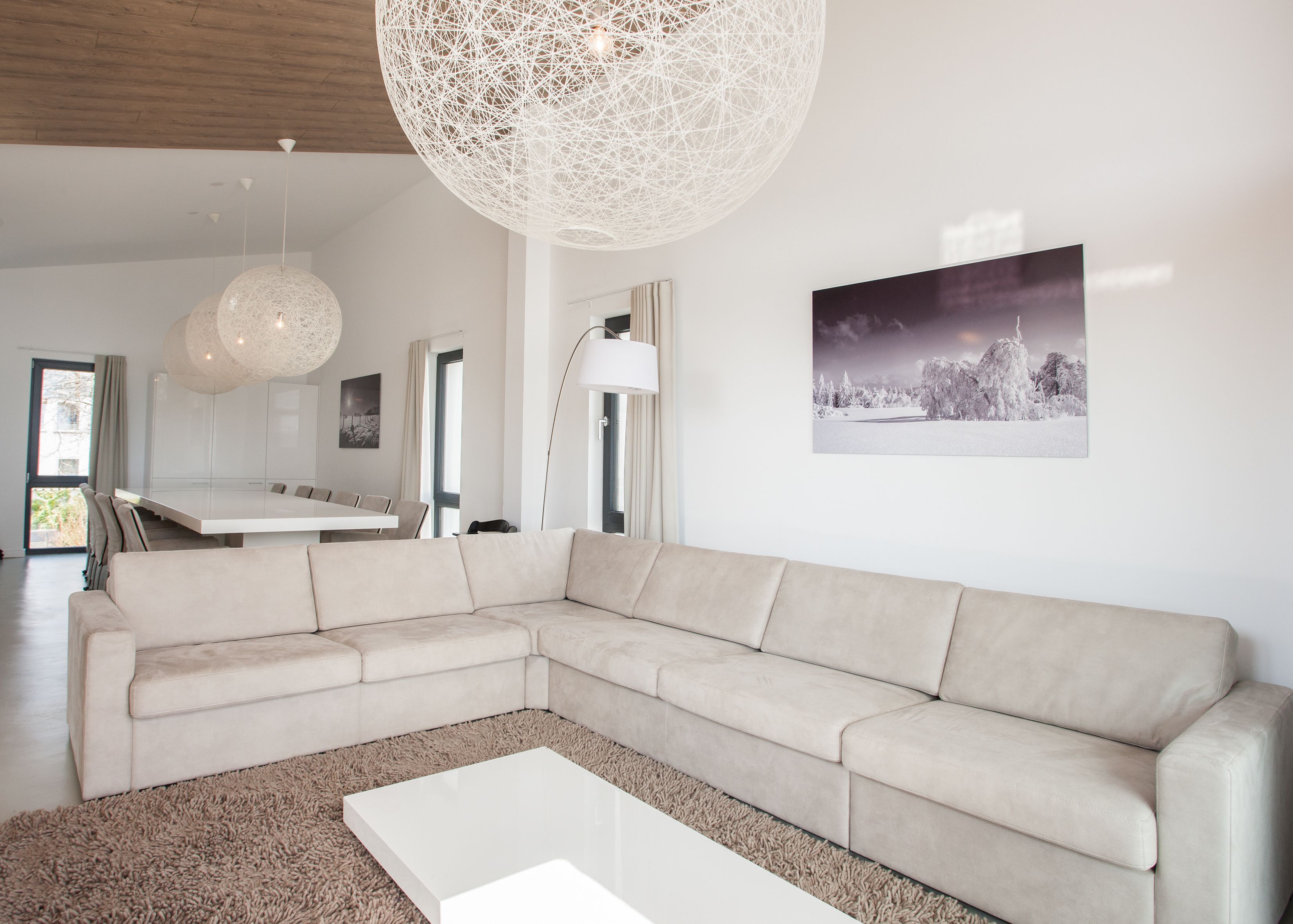 A modern living room with a large L-shaped sectional sofa, a white coffee table, and a large abstract light fixture. There is a dining area with a white table and chairs visible in the background.
