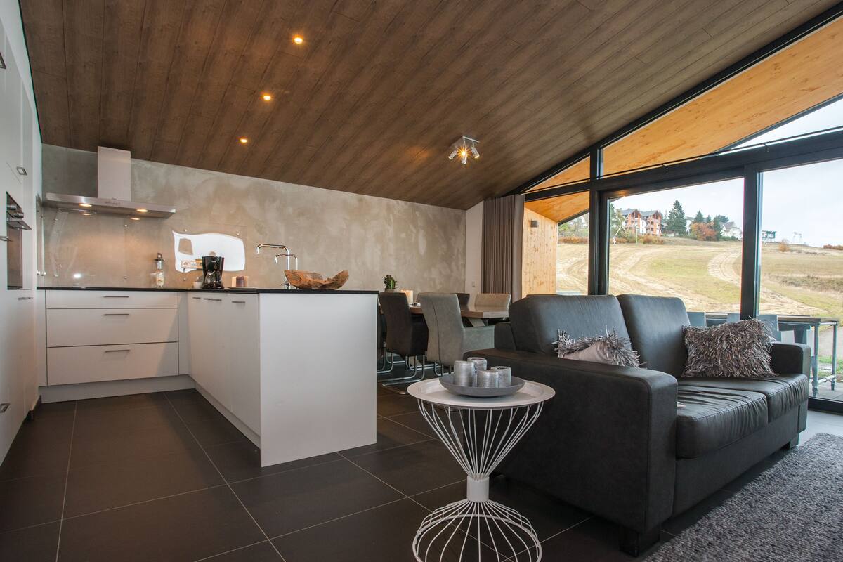 A modern kitchen with a central island, a dining area with a sofa and table, and large windows offering a view of the countryside.