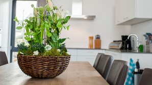 A dining table with a wicker basket containing a floral arrangement, a modern kitchen in the background, and a view of the outdoors through a window. - Villas Winterberg (Winterberg)