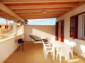 A covered patio with a table and chairs, a lounge chair, and a view of buildings in the background. - VersoSud - Apartment Vicino Lungomare 09 (Ugento)