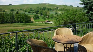 Ein Balkon mit Korbmöbeln und einem Tisch, mit Blick auf eine grüne Landschaft mit sanften Hügeln und einem kleinen Haus in der Ferne.