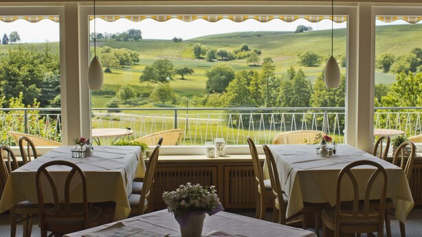 Ein Restaurant mit Tischen und Stühlen, Blick auf grüne Felder und Hügel, sowie hängende Lampen.
