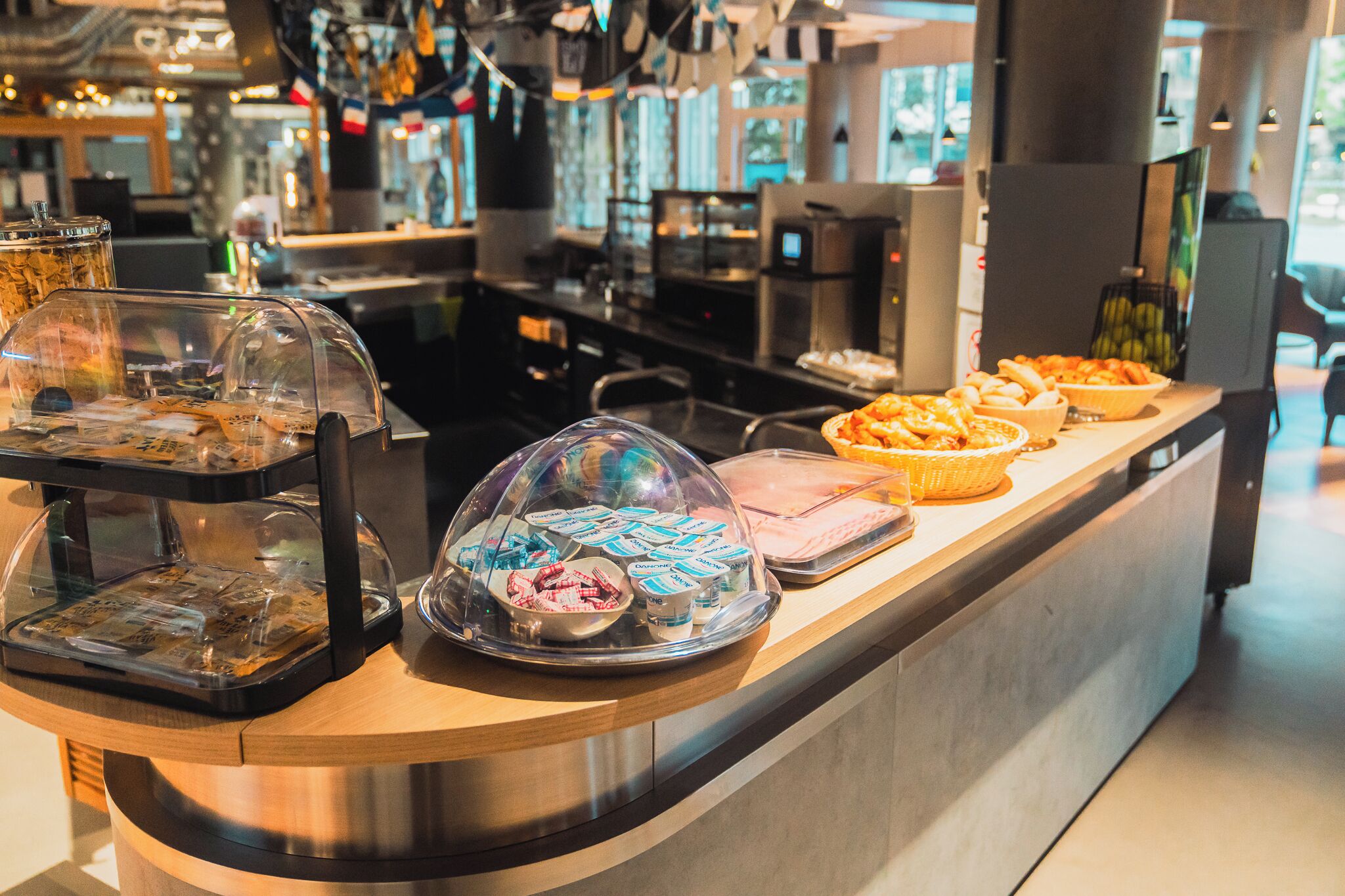 Mostrador de buffet con diversos alimentos cubiertos por campanas de cristal, una máquina de café y un área de estar al fondo.