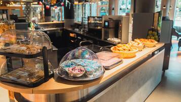 Mostrador de buffet con diversos alimentos cubiertos por campanas de cristal, una máquina de café y un área de estar al fondo.