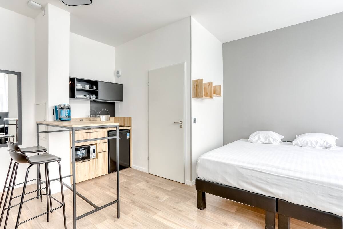 A modern, compact room with a kitchenette, a bed with white bedding, and a small seating area.