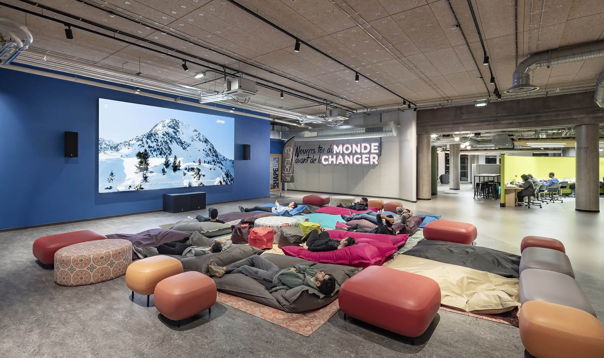 A modern workspace with a large screen displaying a mountain scene, a slogan on the wall, and a group of people lying on colorful cushions.