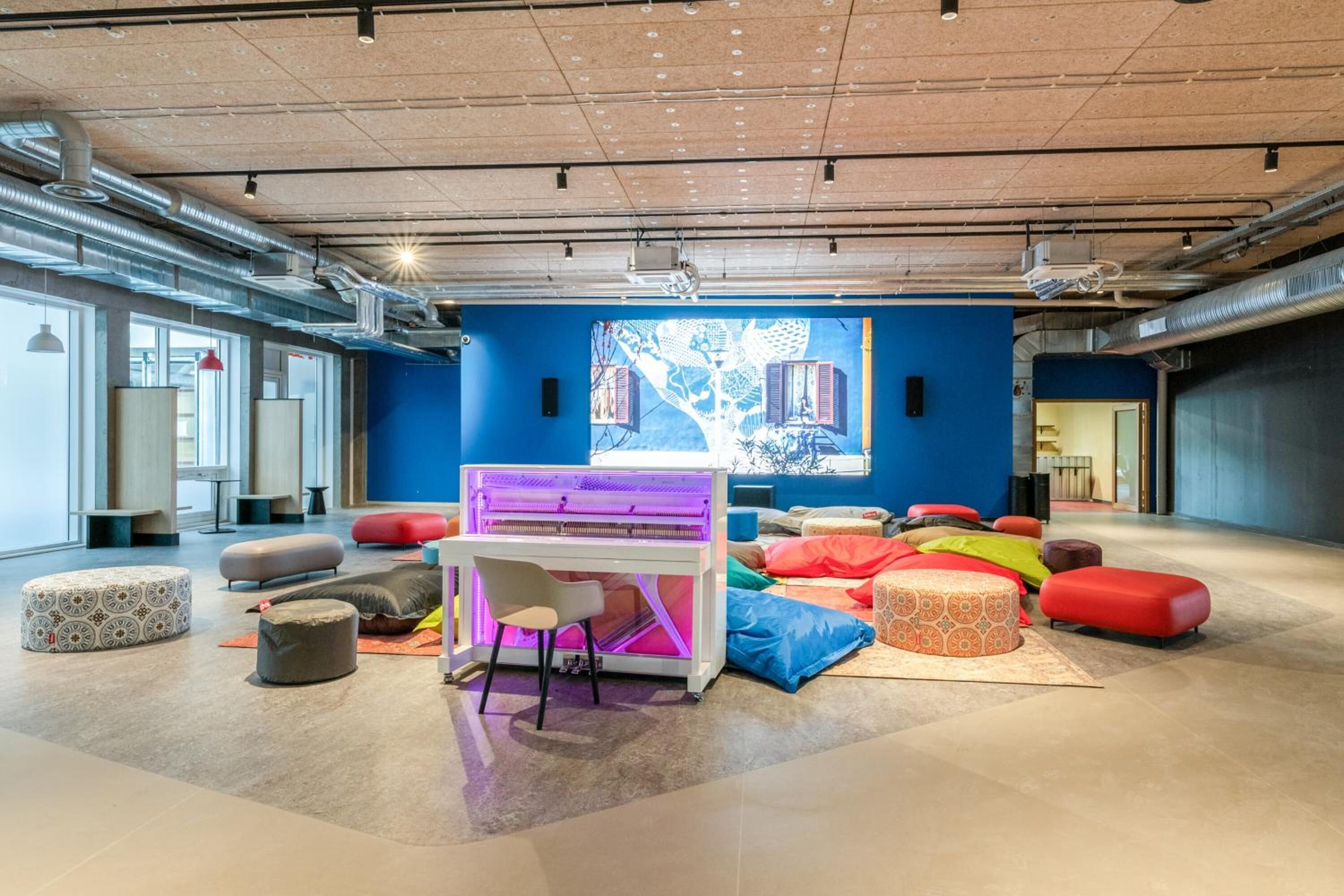 Un espace lounge moderne avec des sièges aux couleurs vives, un piano et une grande fresque murale abstraite.