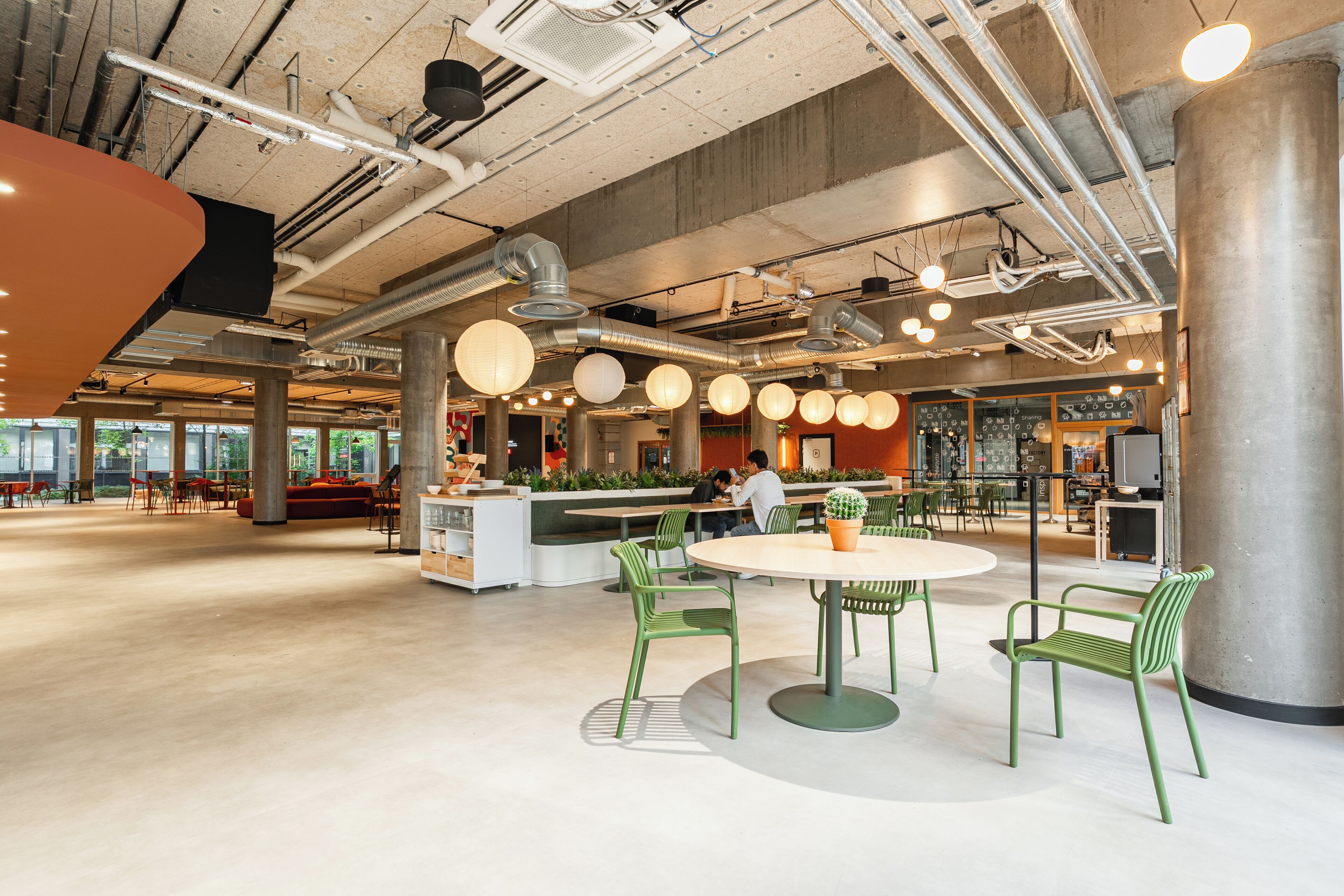 A spacious indoor area with high ceilings, exposed ductwork, and a central seating area with round tables and green chairs.