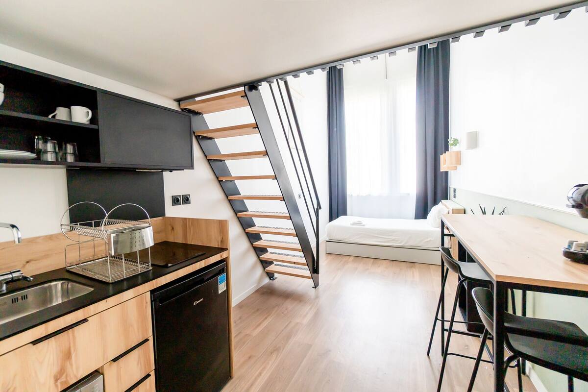A compact kitchen with a built-in dishwasher, a black countertop, and a wooden cabinet. A foldable staircase leads to a sleeping area with a bed and a window with curtains.