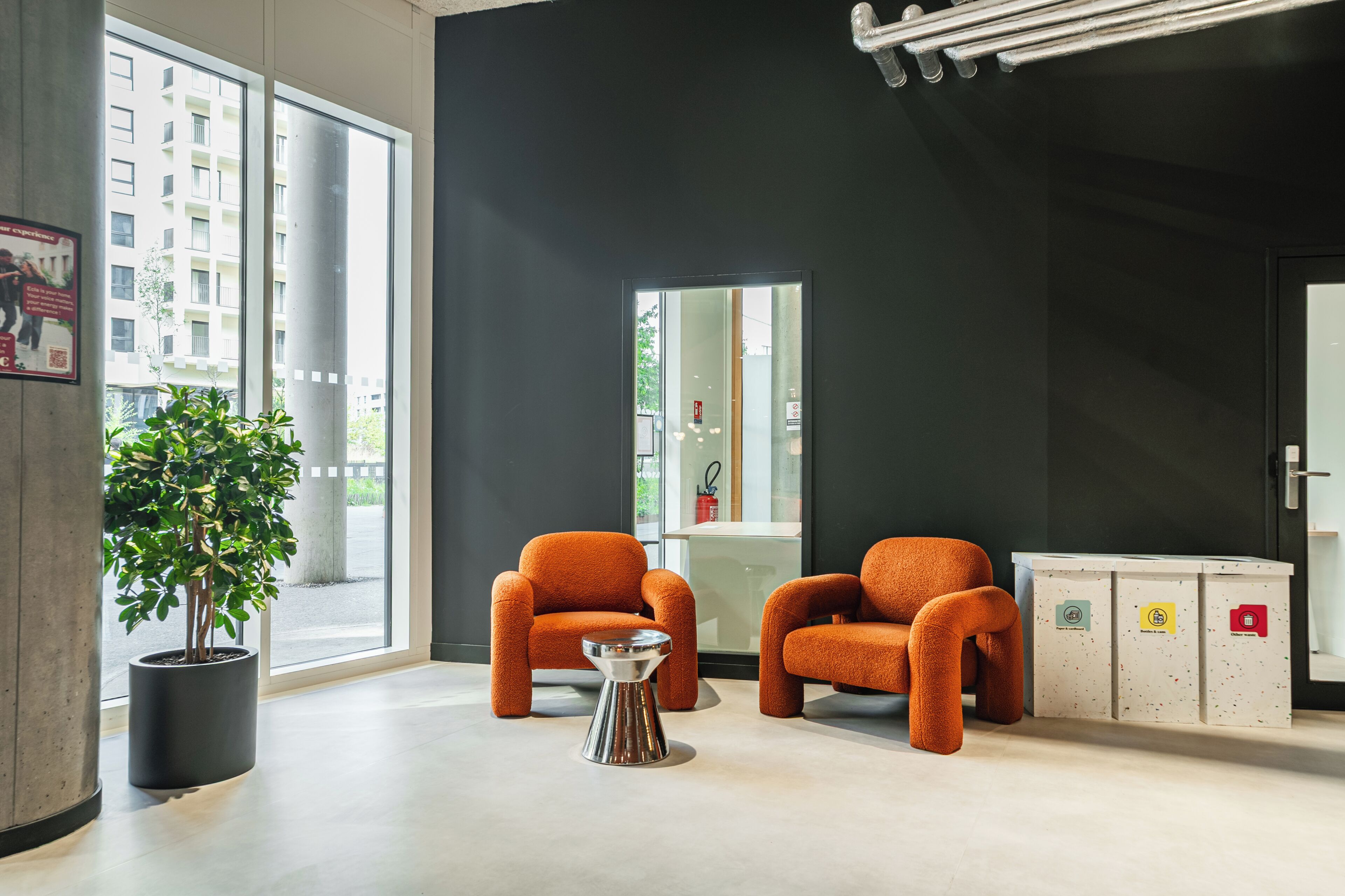 Um lobby moderno com duas poltronas laranja, uma planta em vaso, uma mesinha lateral metálica e uma lixeira com etiquetas de reciclagem.