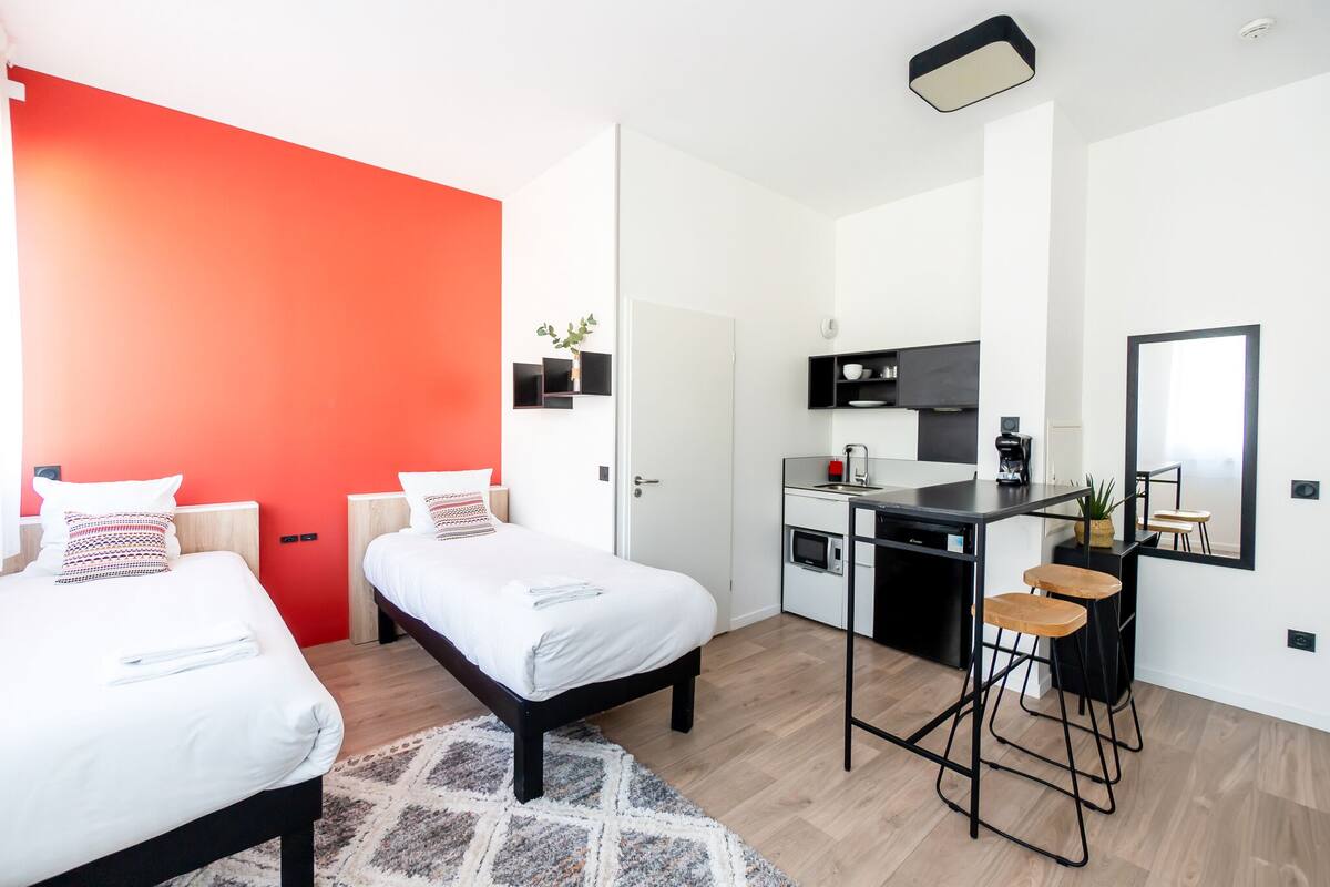 A modern hotel room with two beds, a red accent wall, a kitchenette, and a dining area with bar stools.