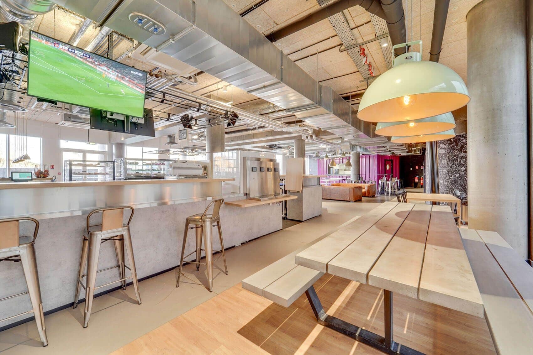 A modern bar area with a long counter, bar stools, and a large TV screen displaying a sports game.
