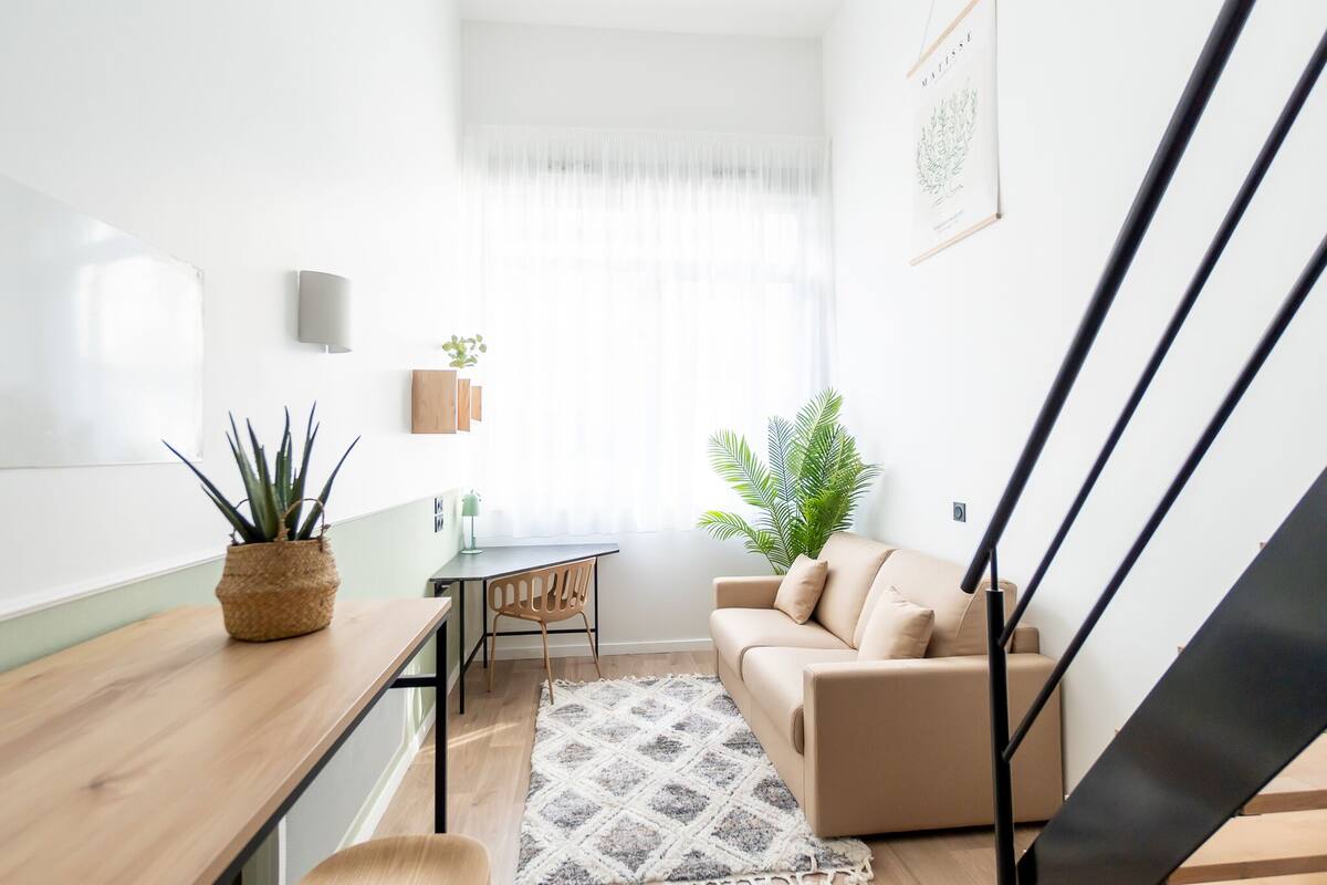 A modern living space with a wooden table, a beige sofa, a small table with a chair, and a potted plant.