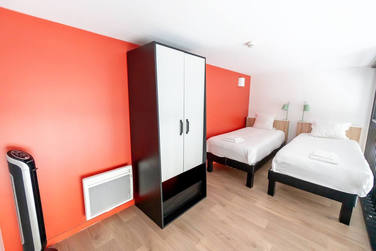 A hotel room with two beds, a wardrobe, a heater, and a small table with lamps.