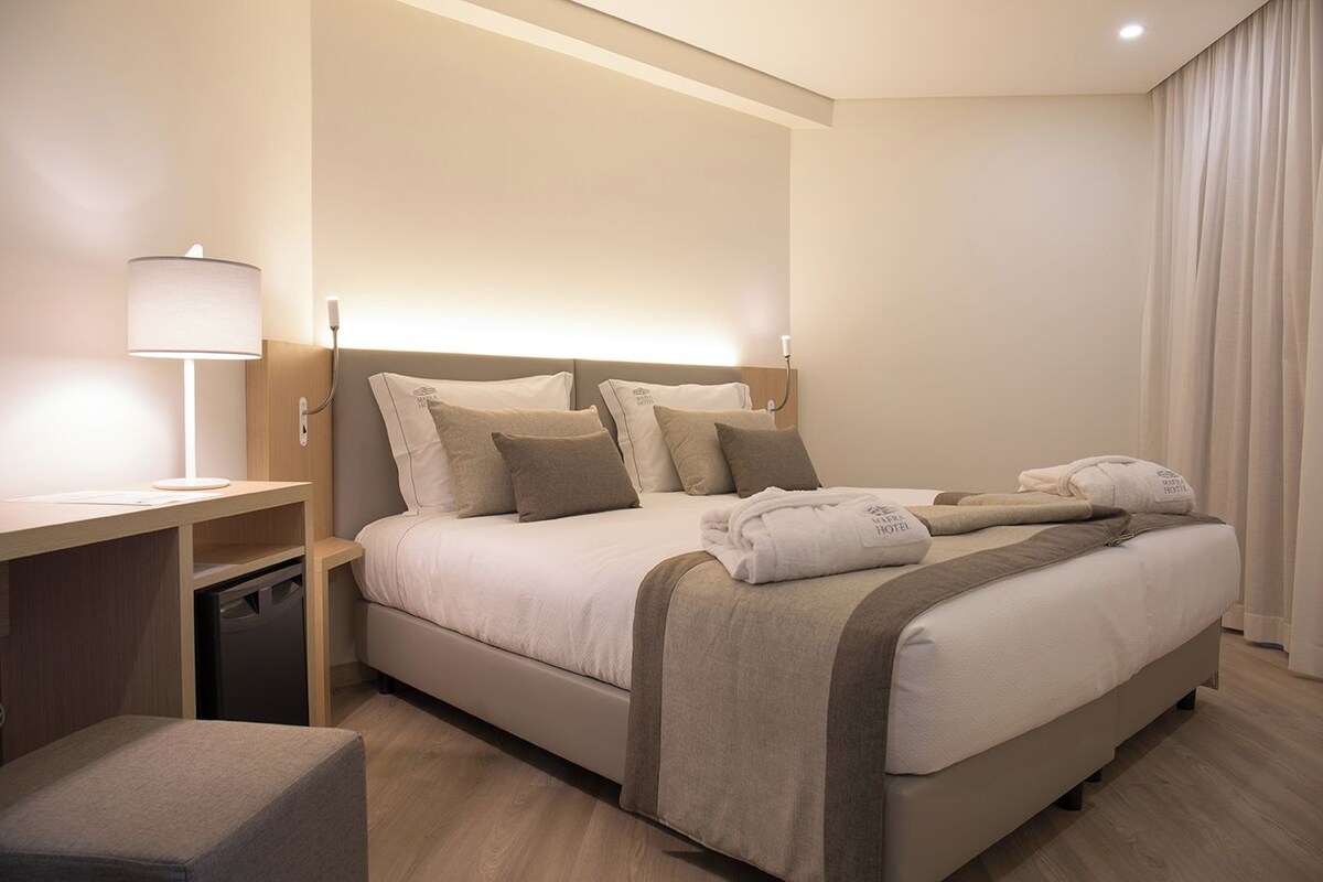 Una habitación de hotel con cama, mesita de noche, lámpara y un pequeño taburete.