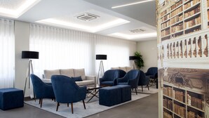 Una sala de estar moderna con un sofá, sillones y una mesa de centro. Hay estanterías a la derecha y un ventanal con cortinas.