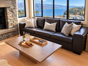 Ein modernes Wohnzimmer mit Kamin aus Stein, einer Ledersofa mit Kissen, einem Couchtisch mit Snacks und Getränken und großen Fenstern mit Panoramablick auf Berge und einen See.