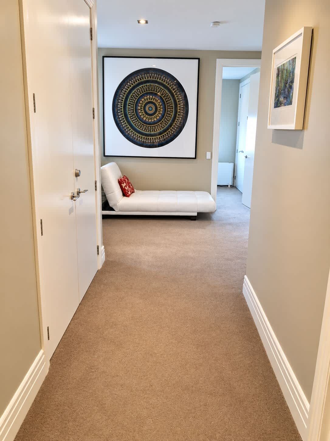 A hallway with a beige carpet, a white sofa with a red pillow, and a large circular artwork on the wall.