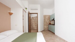 A bedroom with a bed, a green blanket, a white pillow, a wooden door, and a stone wall leading to a kitchen with teal cabinets. - Monolocale del Sole (Morciano di Leuca)