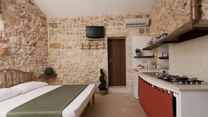 A compact room with a bed, a small kitchenette, a stone wall, and a television. - Monolocale del Sole (Morciano di Leuca)