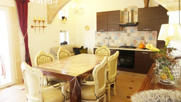 A dining area with a marble table, white chairs, a chandelier, and a kitchen with dark cabinets and a tiled backsplash. - VersoSud - Casa Torre San Giovanni 17 (Ugento)