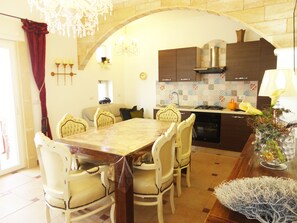 Un espace repas comprenant une table en marbre, des chaises blanches, un lustre et une cuisine avec des meubles sombres et un dosseret carrelé.