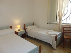 A hotel room with two single beds, a nightstand with a lamp, a chair, and a window with curtains. - VersoSud - Casa Vacanze De Revel 18 (Ugento)