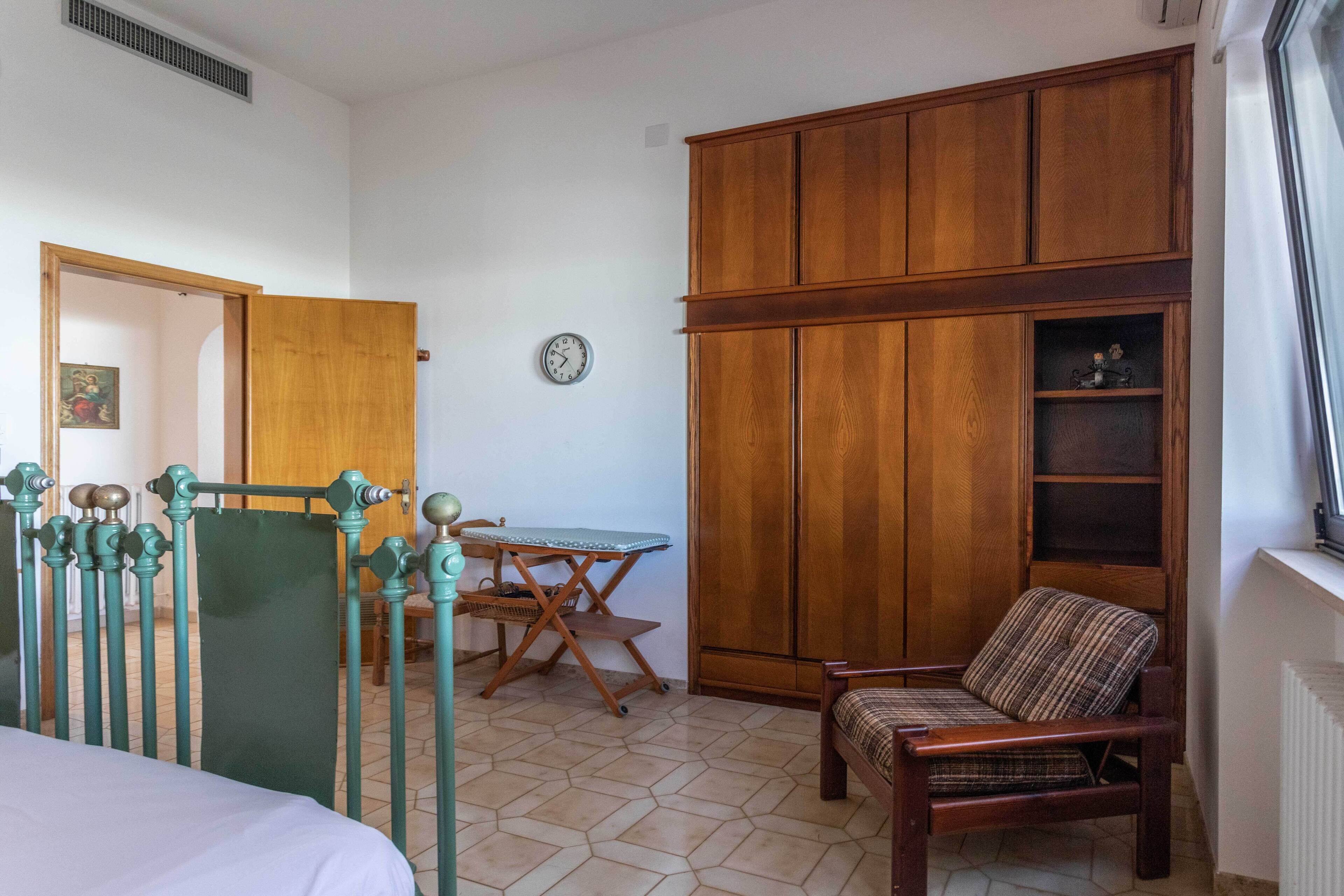 A bedroom with a bed, a wooden wardrobe, a chair, a table with an ironing board, and a clock on the wall.