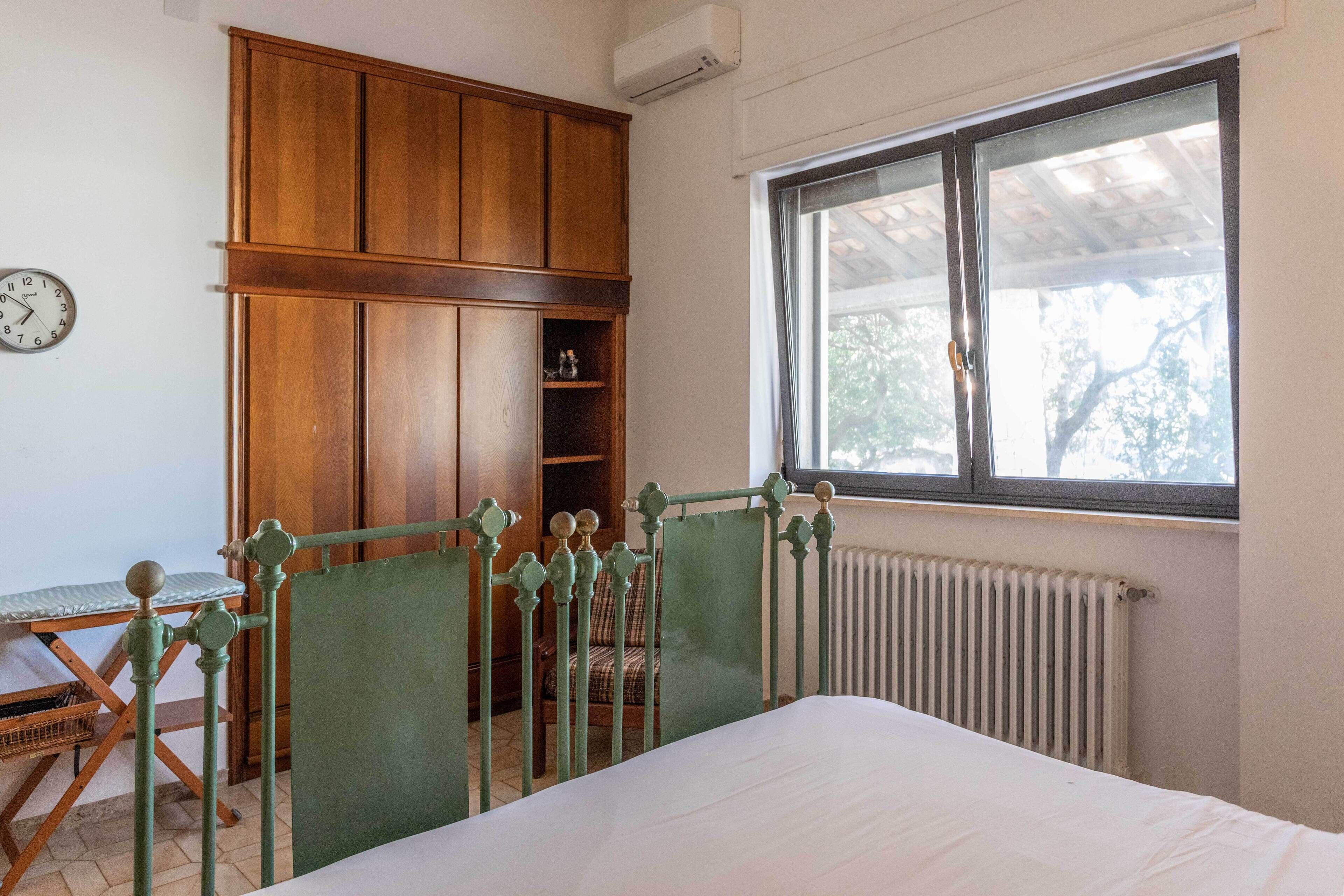 Un dormitorio con cama, armario de madera, radiador, ventana y un reloj en la pared.