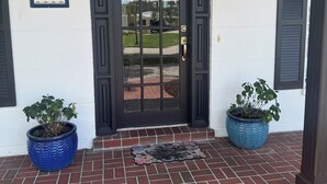 Une maison avec une porte d’entrée noire, une plaque de numéro, deux plantes en pot et un porche en briques.