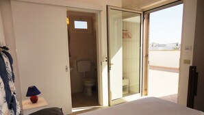 Un dormitorio con cama, una mesita de noche con lámpara y un baño visible a través de una puerta abierta.