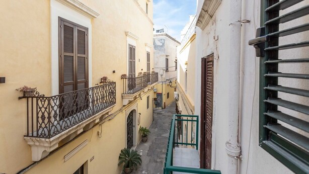 A narrow alleyway between buildings with balconies, a small garden area, and a clear sky. - Palazzo Raheli (Gallipoli)
