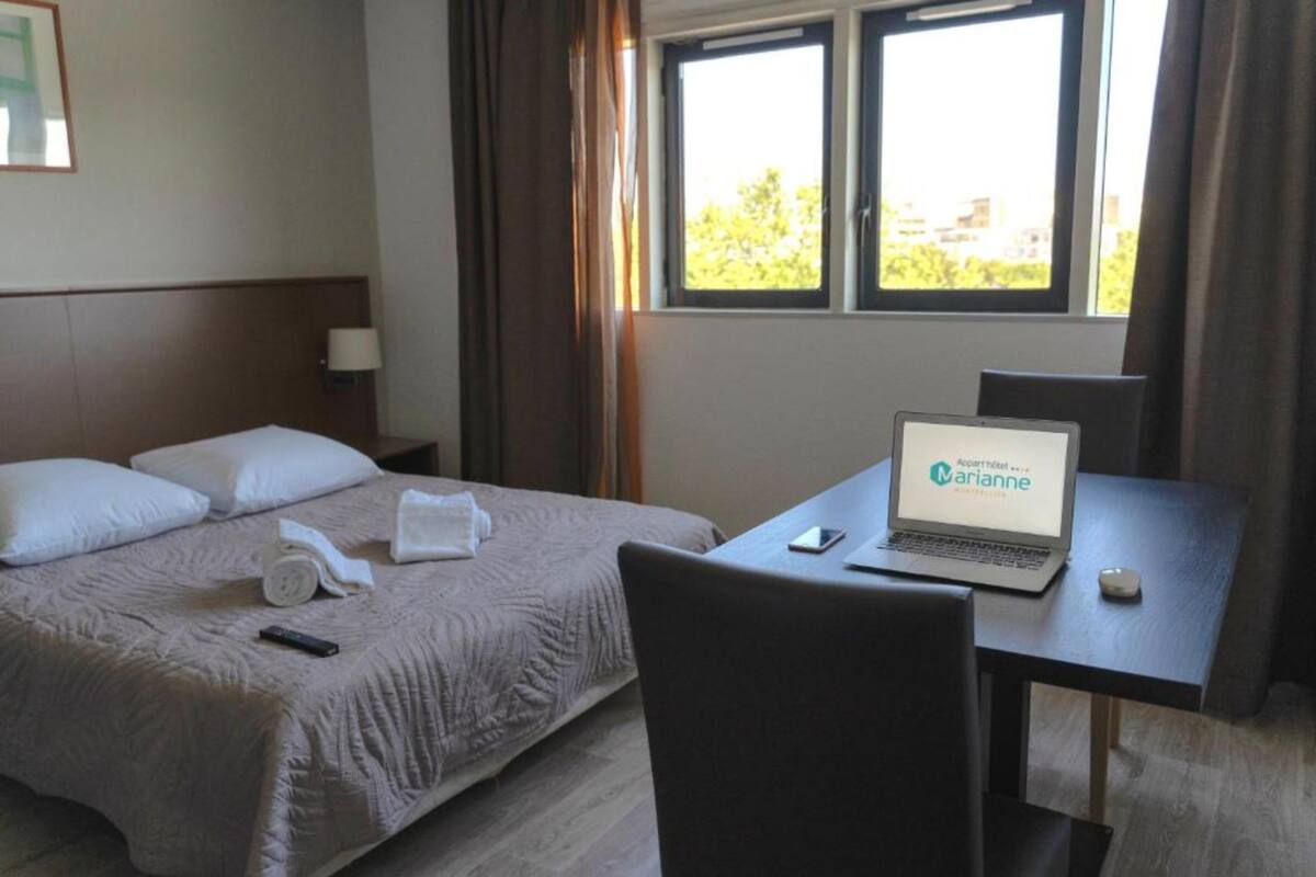 Une chambre d’hôtel avec un lit, un bureau avec un ordinateur portable, une chaise et une fenêtre avec des rideaux.